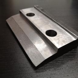 M-TECH Sp. Z o.o. - Metalowy element z dwoma otworami, obrobiony z widocznymi śladami frezowania, leżący na ciemnym, drewnianym blacie.