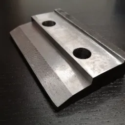 Metalowy element z dwoma otworami, obrobiony z widocznymi śladami frezowania, leżący na ciemnym, drewnianym blacie.