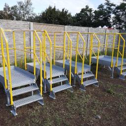 M-TECH Sp. Z o.o. - Metalowe schody przemysłowe z żółtymi poręczami, zabezpieczone folią, ustawione na zewnątrz na ziemi, z betonowym ogrodzeniem w tle.