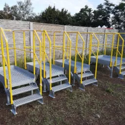 Metalowe schody przemysłowe z żółtymi poręczami, zabezpieczone folią, ustawione na zewnątrz na ziemi, z betonowym ogrodzeniem w tle.