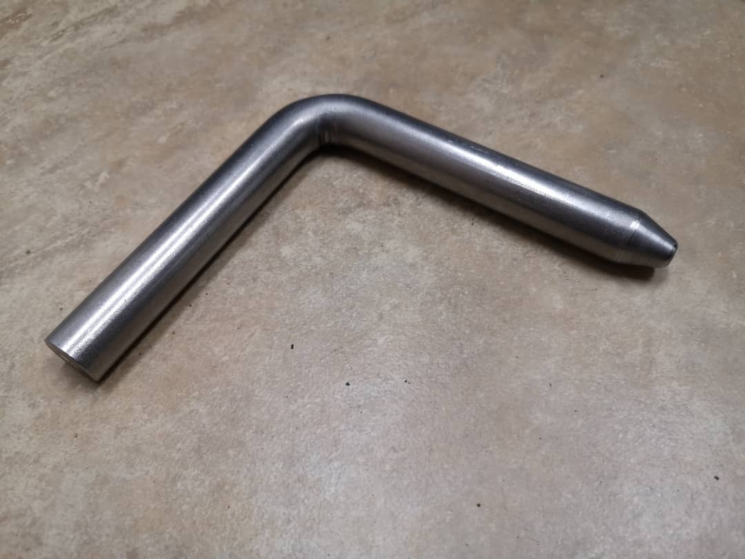 Metalowy pręt zagięty pod kątem prostym, z lekko stożkowym zakończeniem, leżący na szarej powierzchni.