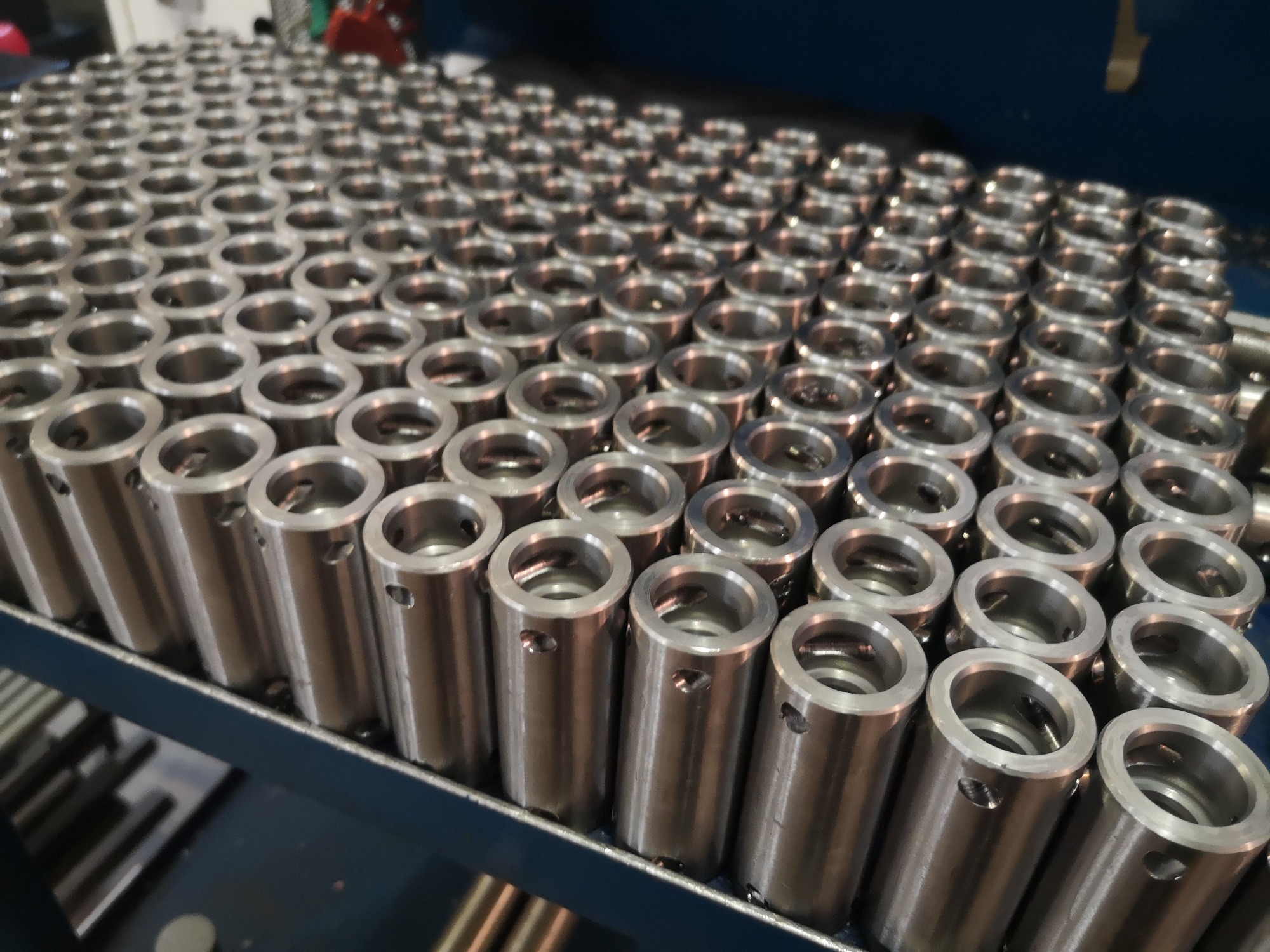 Rząd cylindrycznych, metalowych elementów z otworem wewnątrz, ułożonych na metalowej półce, widoczne otwory boczne w każdym elemencie.