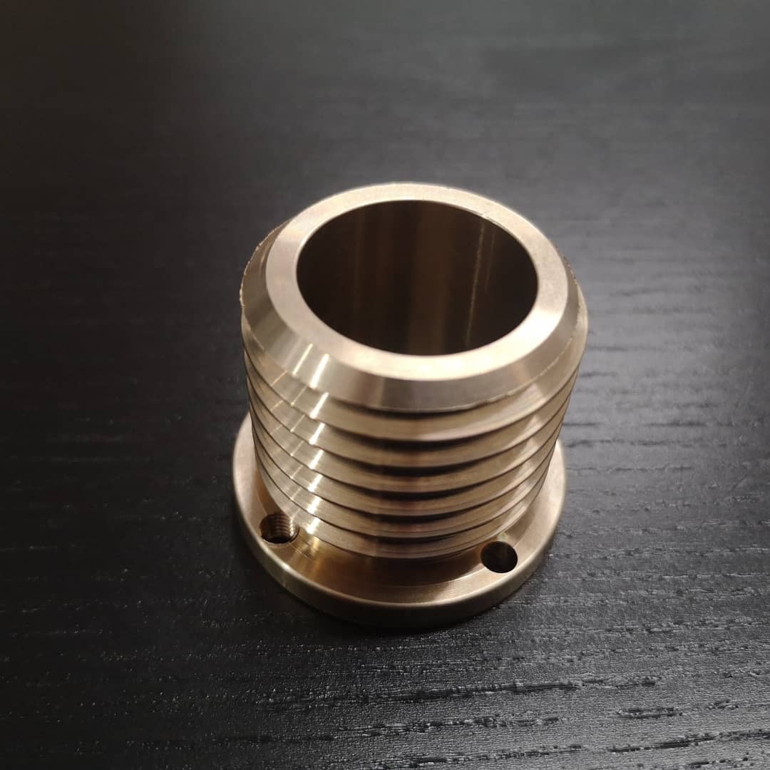 Mosiężny element gwintowany z kołnierzem, widoczne otwory montażowe, leżący na ciemnym, drewnianym blacie.