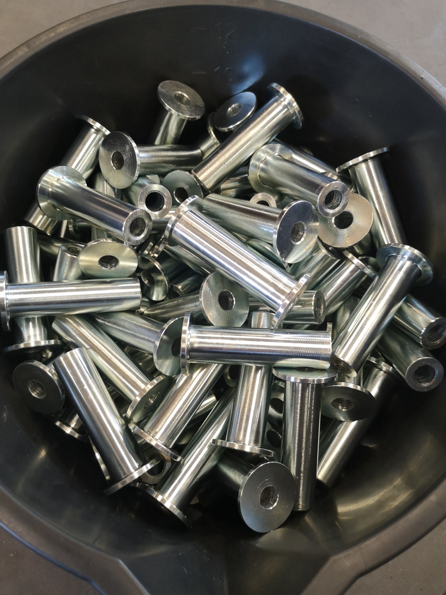Partia obrobionych, metalowych tulejek z kołnierzem i otworem, zgromadzona w plastikowym pojemniku, widoczne ślady obróbki skrawaniem.