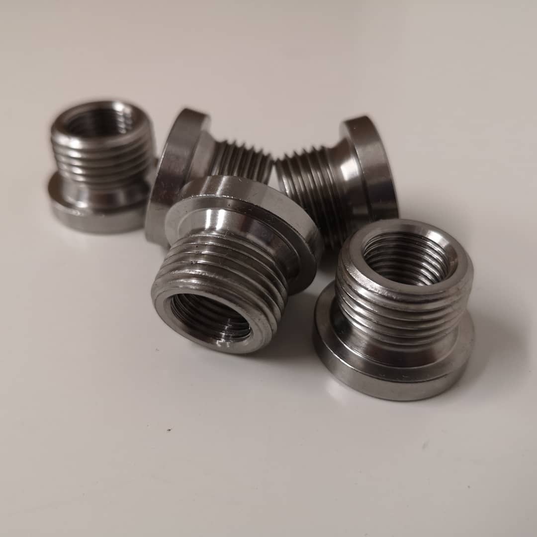 Srebrne, metalowe tuleje z gwintem wewnętrznym i zewnętrznym, ułożone na białym tle, widoczne ślady obróbki.