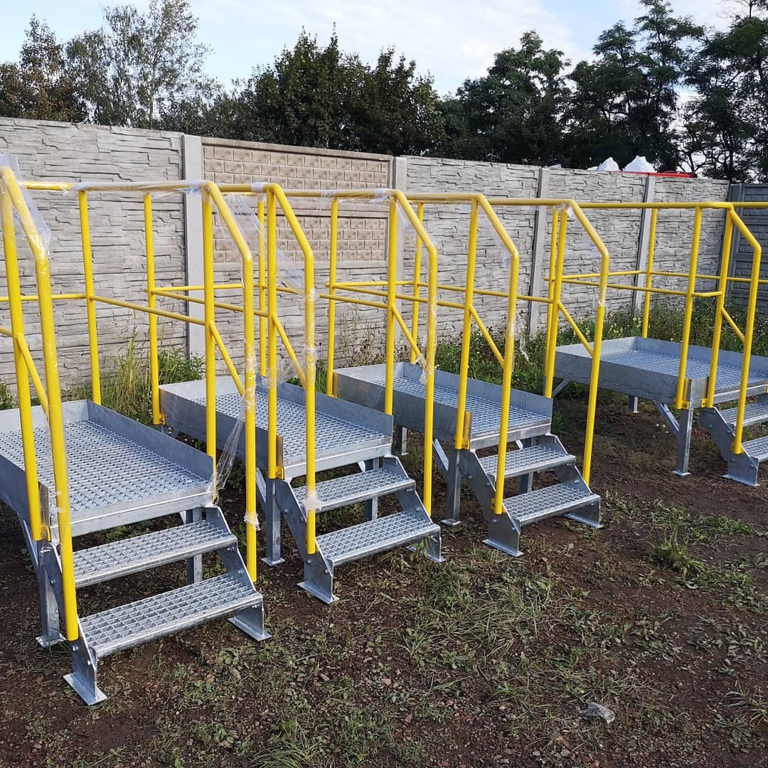 Metalowe schody przemysłowe z żółtymi poręczami, zabezpieczone folią, ustawione na zewnątrz na ziemi, z betonowym ogrodzeniem w tle.