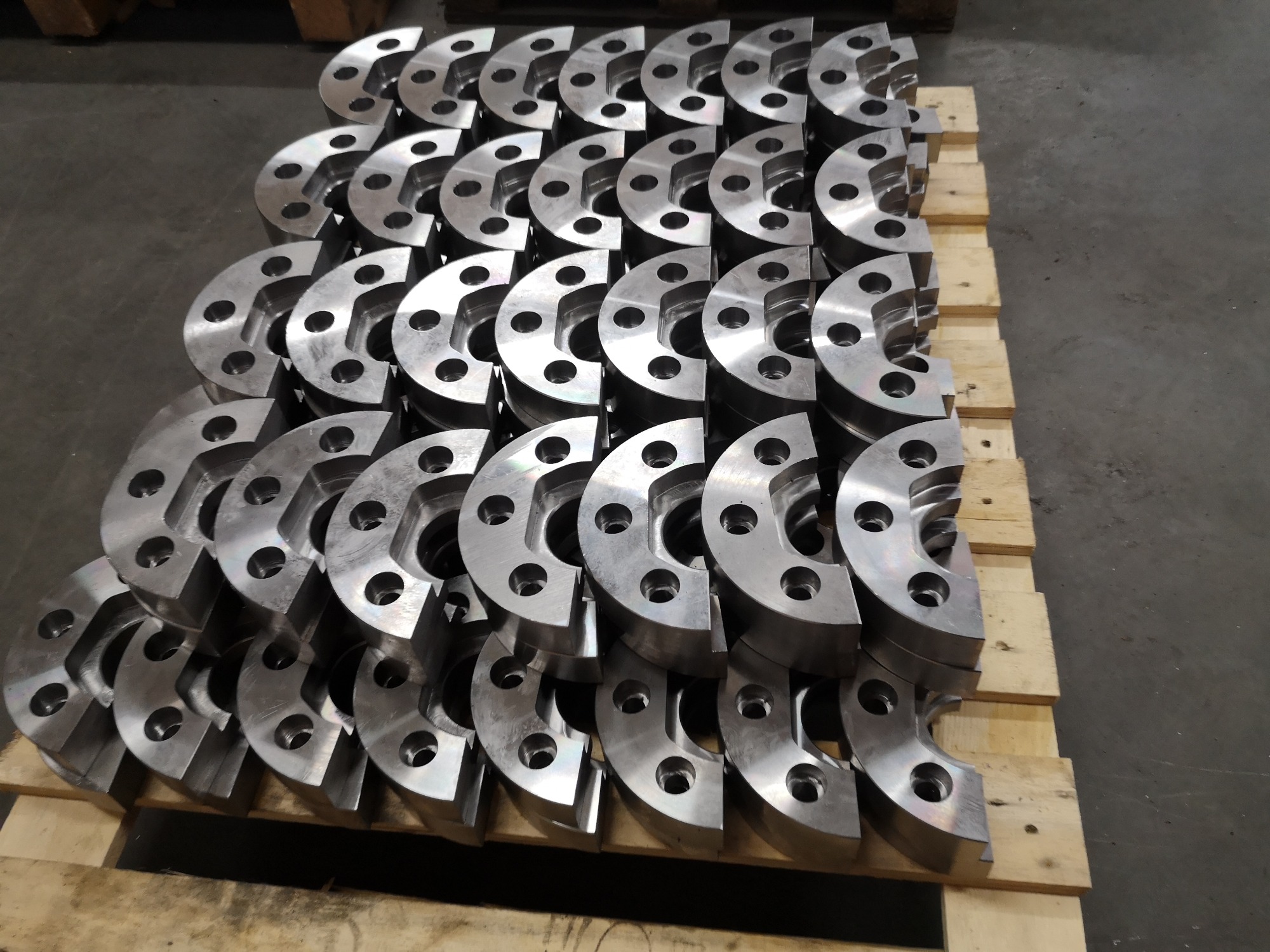 Stos obrobionych CNC, metalowych flansz z otworami, ułożonych równo na drewnianej palecie w hali produkcyjnej.