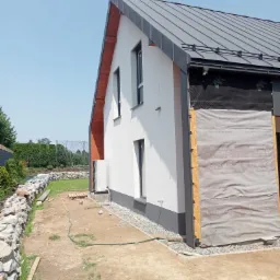 Dom jednorodzinny w trakcie prac elewacyjnych: fragment ściany z widoczną warstwą izolacyjną zabezpieczoną membraną, fragment dachu z blachy na rąbek stojący, biała elewacja, szara podmurówka...