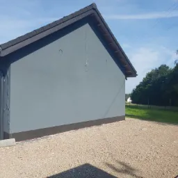 Świeżo otynkowana szara elewacja budynku gospodarczego z czarnym dachem i orynnowaniem; widoczne przewody wystające ze ściany i podłoże z kamyczków.