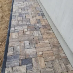 Ułożona ścieżka z kostki brukowej w odcieniach brązu i szarości, przylegająca do jasnej ściany budynku, z widoczną czarną obwódką i fragmentem ziemi obok.