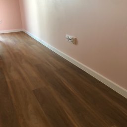 RAF-BUD Profesionalnie Wykladziny - Ukończona podłoga z paneli imitujących drewno w pokoju z różowymi ścianami, widoczne listwy przypodłogowe i gniazdka elektryczne.