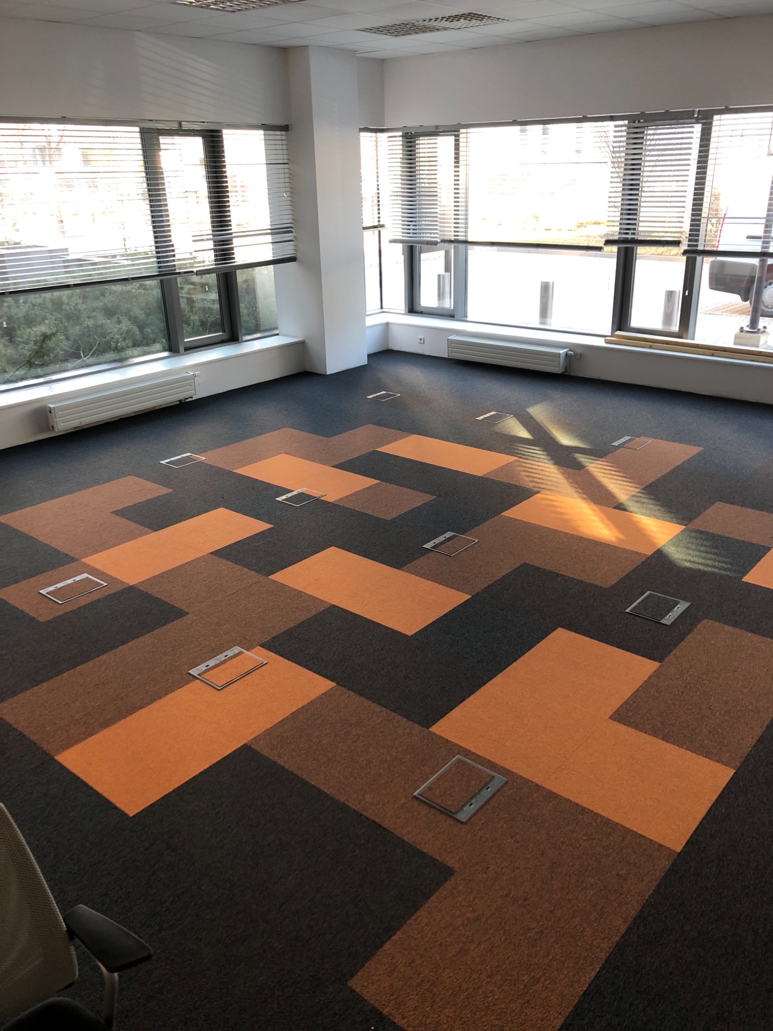 Wykładzina dywanowa w biurze w geometryczny wzór z widocznymi puszkami podłogowymi, oświetlona promieniami słońca wpadającymi przez żaluzje.