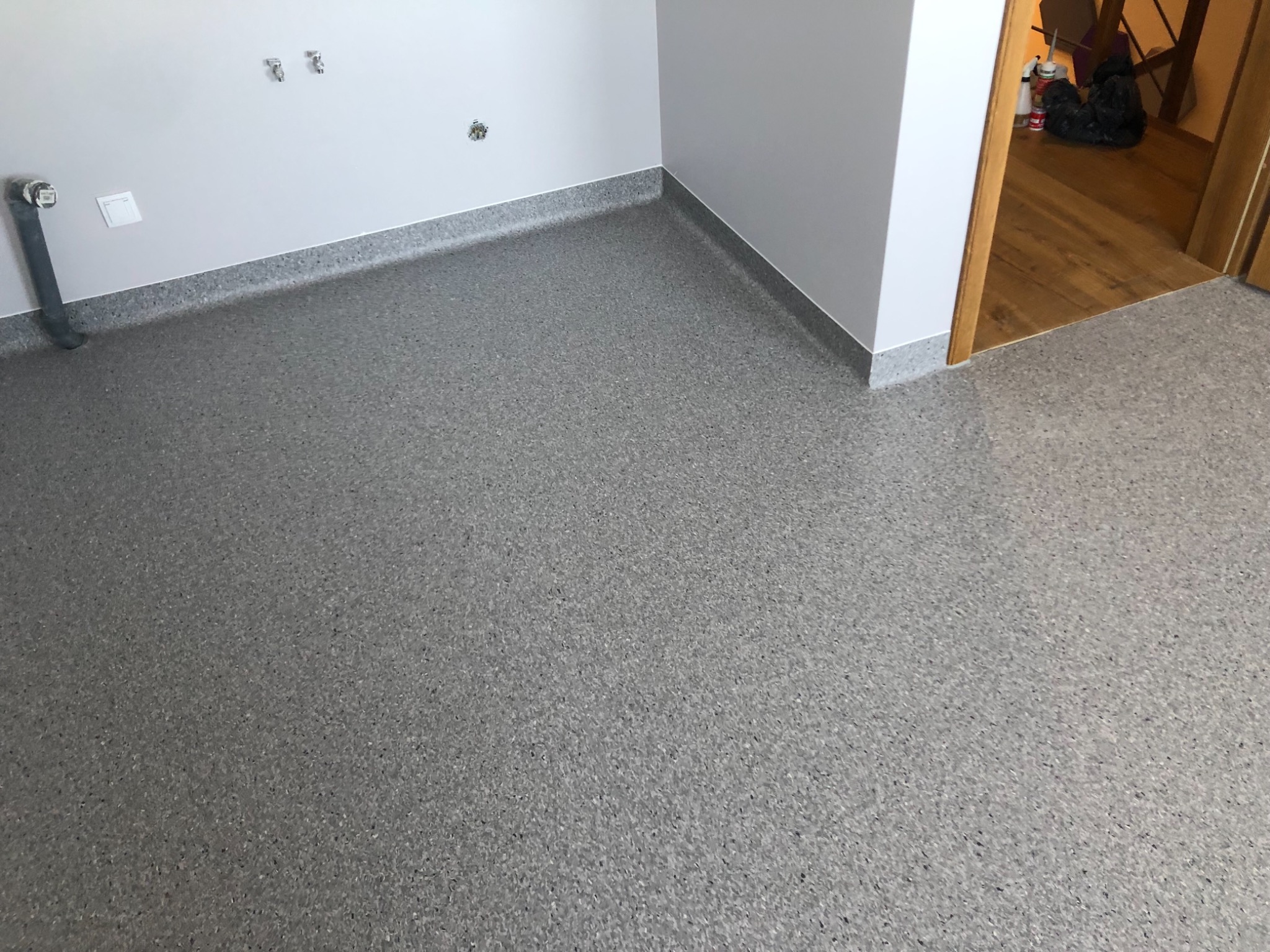 Szara, nakrapiana podłoga z wykładziny PCV w rogu pomieszczenia, widoczne listwy przypodłogowe i instalacja wodna na ścianie.