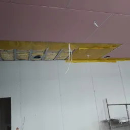 Montaż sufitu podwieszanego: widoczne metalowe profile, izolacja z folią, otwory na oświetlenie i częściowo zamontowane płyty kartonowo-gipsowe w kolorze różowym.