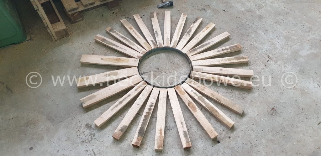 Drewniane listewki ułożone promieniście wokół metalowego okręgu, tworzące dekoracyjny element w warsztacie.