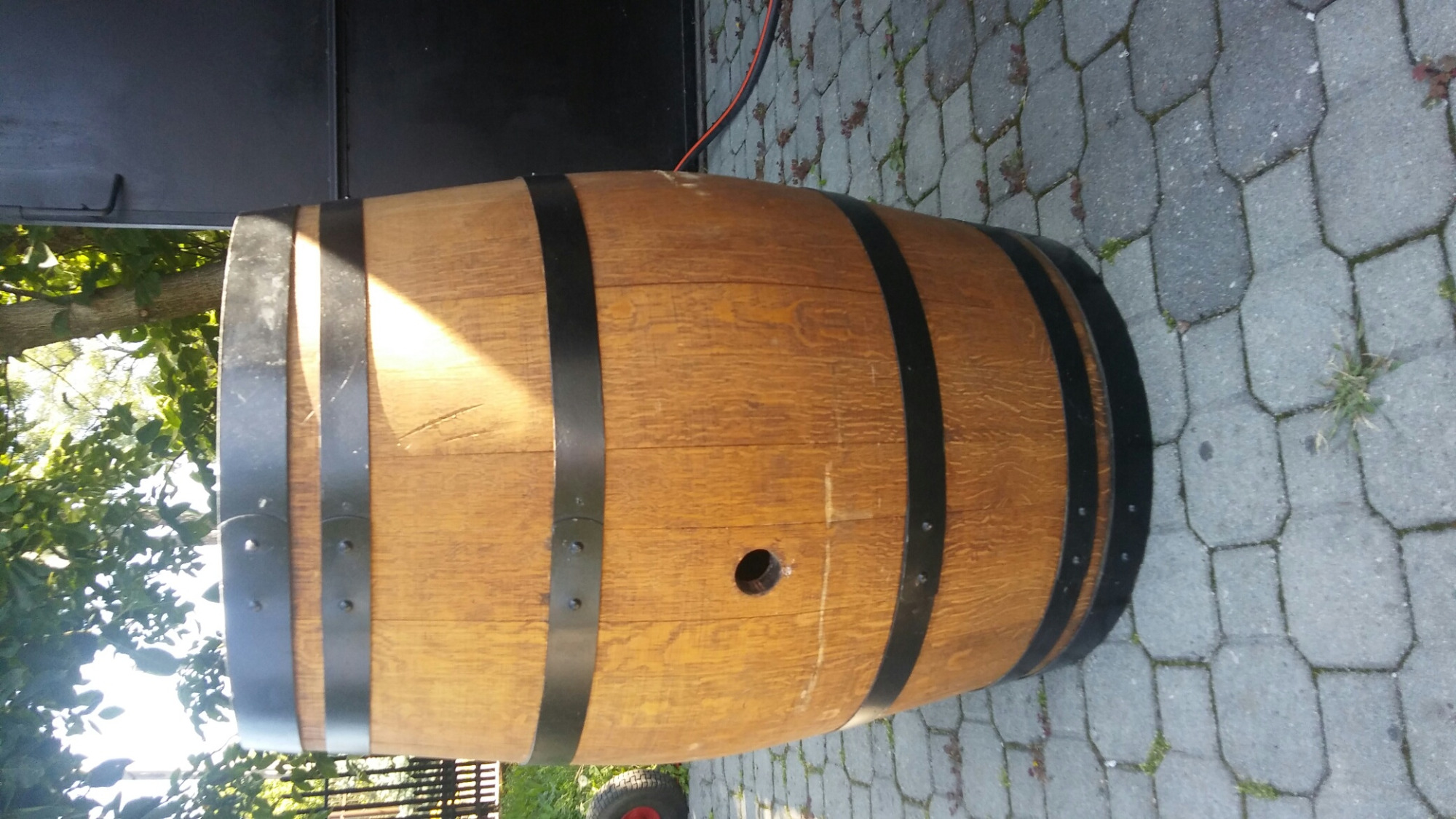 Drewniana beczka z czarnymi obręczami leżąca na szarym bruku, widoczny otwór w boku beczki, drzewo w tle.