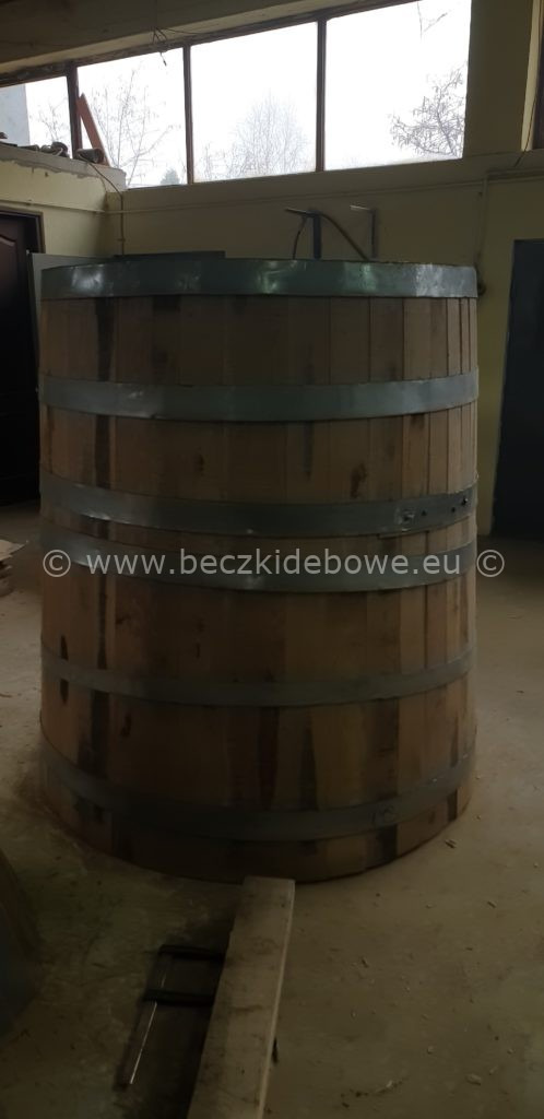 Duża, drewniana beczka z metalowymi obręczami w warsztatowym otoczeniu, widoczne okno z drzewami w tle.