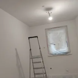 Świeżo pomalowane na biało wnętrze pokoju z zabezpieczonym folią oknem i aluminiową drabiną, widoczne ślady farby na podłodze.