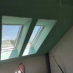 Dwa okna dachowe w trakcie montażu w poddaszu wykończonym płytami gipsowo-kartonowymi, widoczne przewody instalacyjne i rury kanalizacyjne.