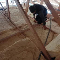 Pracownik w czarnym kombinezonie instaluje izolację z wełny mineralnej na poddaszu, widoczne drewniane krokwie i fragment przewodu wentylacyjnego.