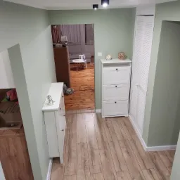 Korytarz w odcieniach zieleni i brązu z drewnianą podłogą i białymi meblami, widok na salon z małym stolikiem i krzesłem dla dziecka.