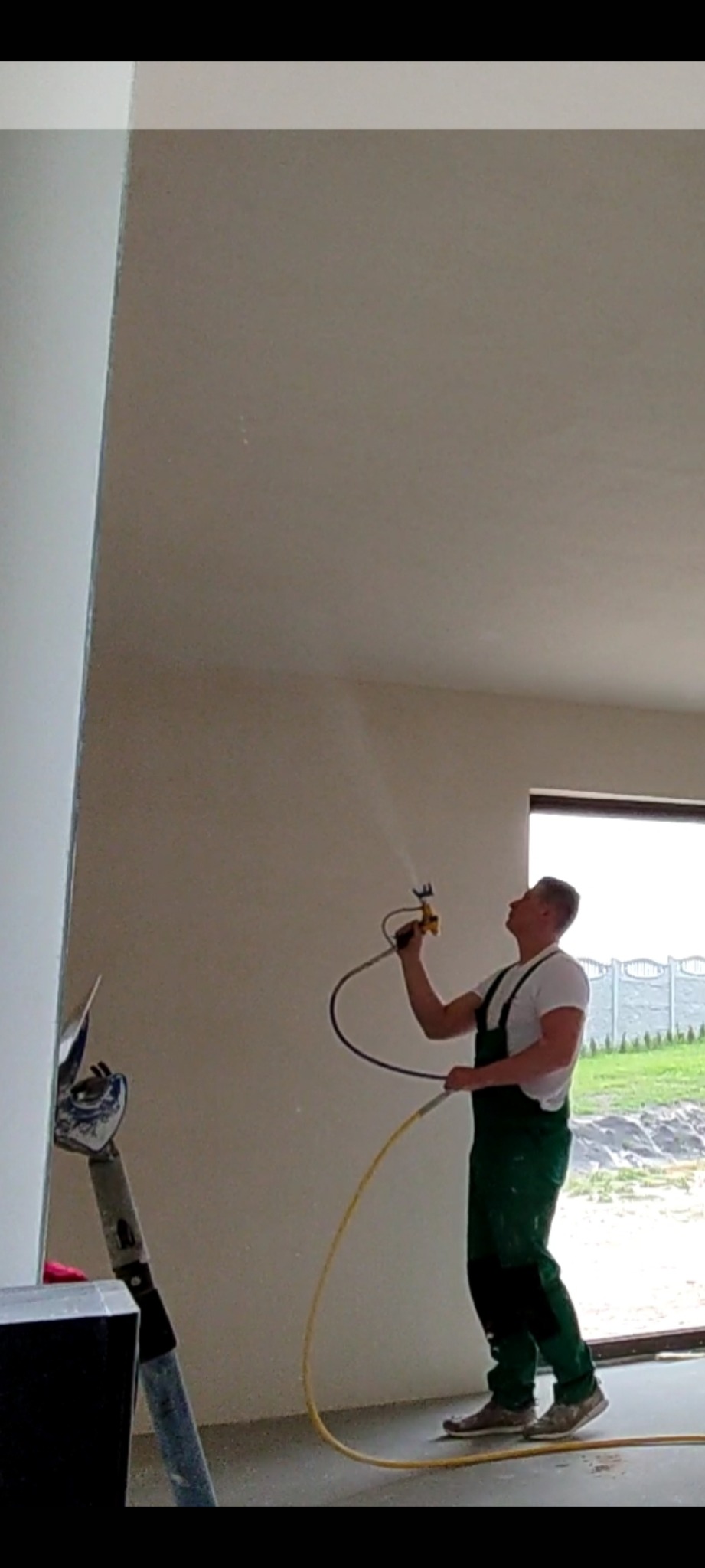 Pracownik w zielonym kombinezonie natryskowo aplikuje gładź na ścianę w nowym budynku, widoczny szpachel i okno z widokiem na zewnątrz.