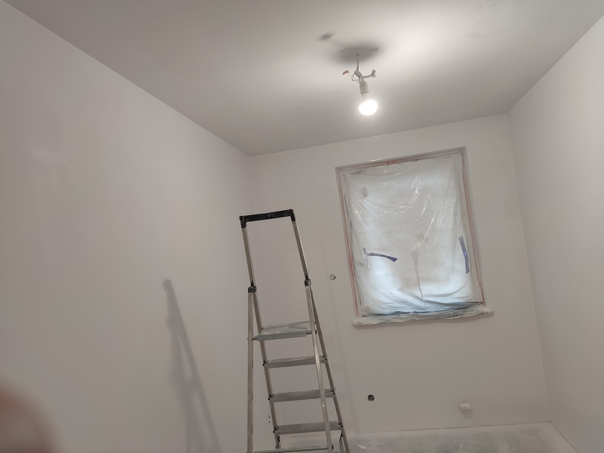 Świeżo pomalowane na biało wnętrze pokoju z zabezpieczonym folią oknem i aluminiową drabiną, widoczne ślady farby na podłodze.