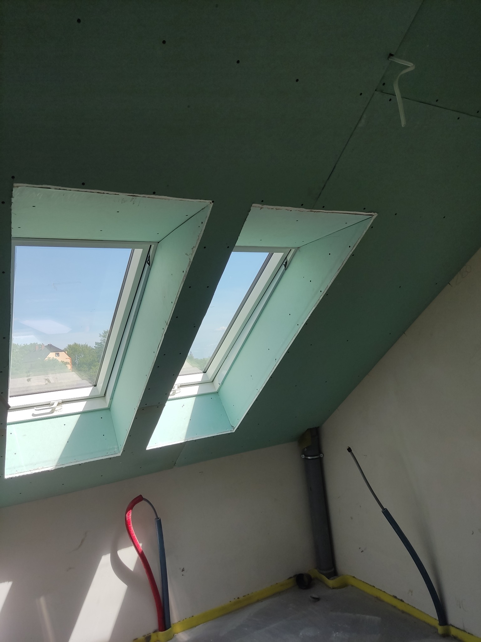 Dwa okna dachowe w trakcie montażu w poddaszu wykończonym płytami gipsowo-kartonowymi, widoczne przewody instalacyjne i rury kanalizacyjne.