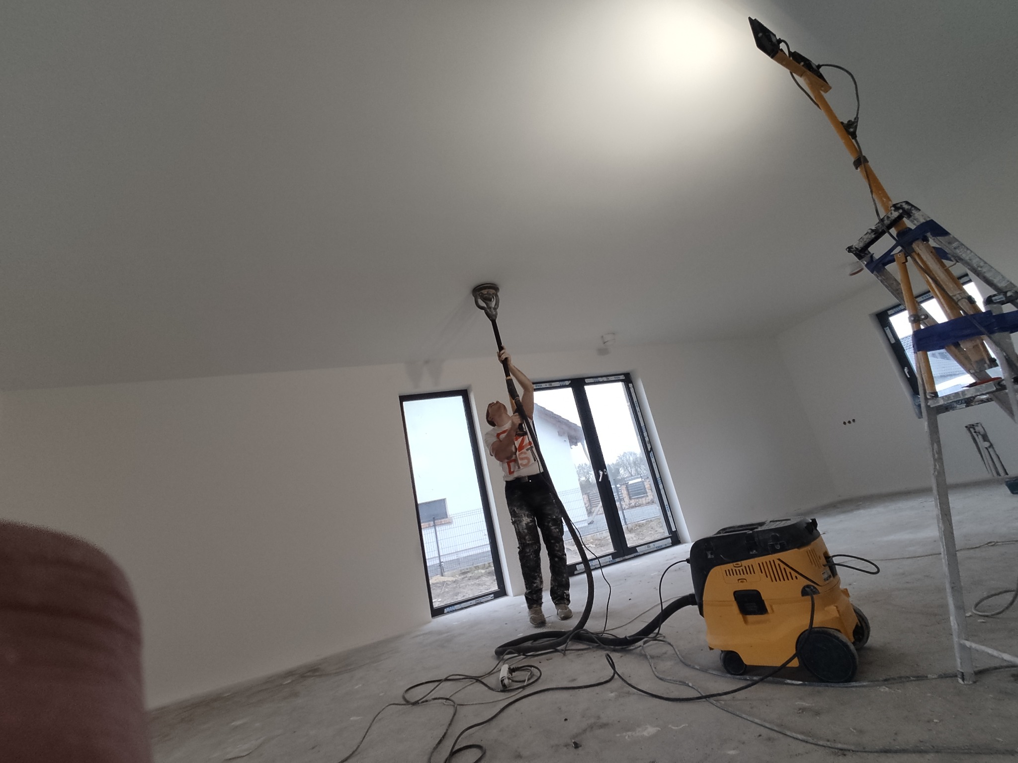 Pracownik szlifuje sufit za pomocą szlifierki teleskopowej podłączonej do odkurzacza przemysłowego w jasnym, nowo wykończonym pomieszczeniu z dużymi oknami.