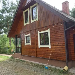 Drewniany dom jednorodzinny z brązową elewacją po renowacji, widoczne odświeżone ramy okienne i dekoracyjne lampki, na ziemi stoi miotła.