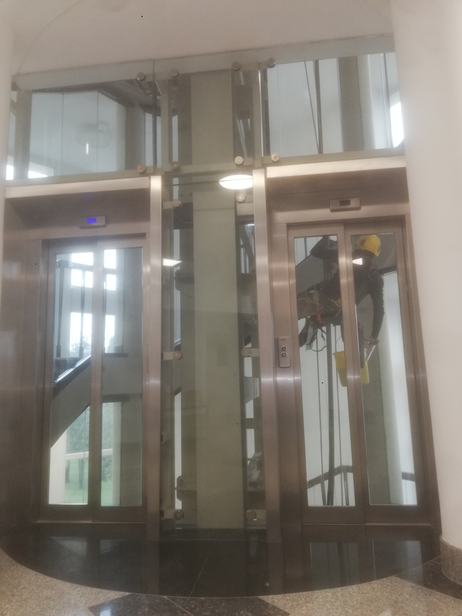 Pracownik w uprzęży i kasku myje szklane ściany szybu windy wewnątrz budynku, widoczne odbicie schodów w drugiej windzie.