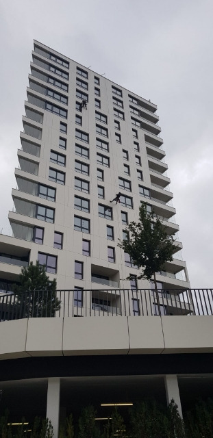 Pracownicy na linach wykonują prace na wysokościach na elewacji wysokiego budynku mieszkalnego w Grabowie nad Pilicą, widok z dołu na tle pochmurnego nieba.