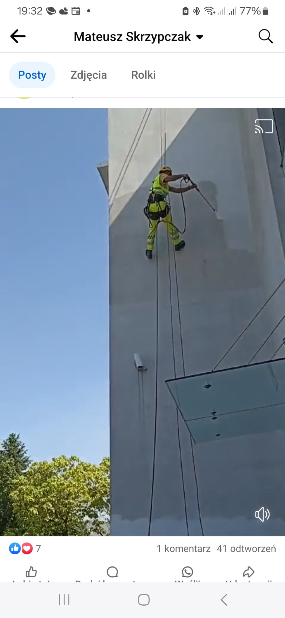 Pracownik w żółtym kombinezonie, zawieszony na linach, myje elewację wysokiego budynku za pomocą myjki ciśnieniowej.