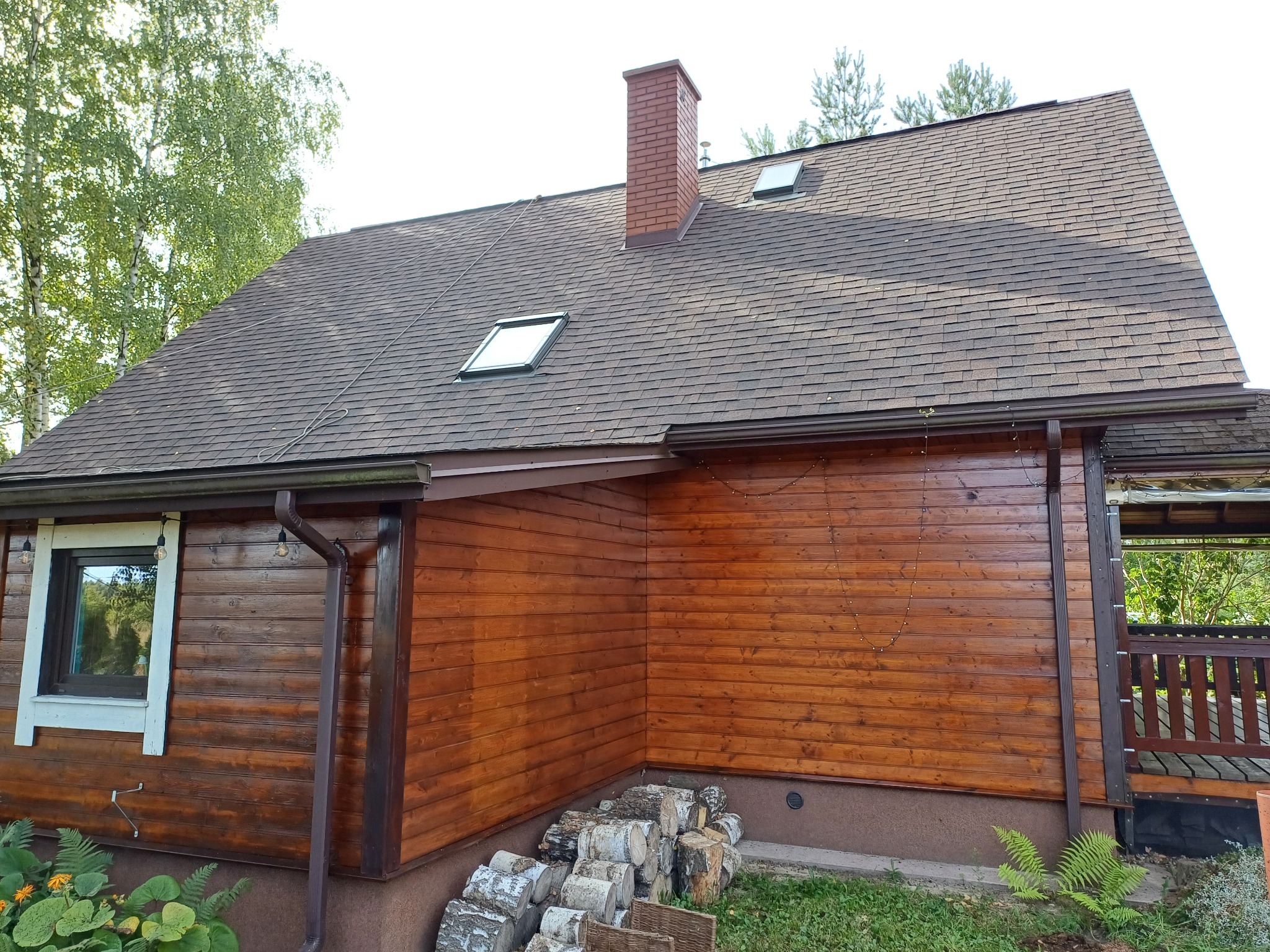 Dom z drewnianą elewacją pomalowaną na brązowo, z brązowym dachem pokrytym gontem bitumicznym, ceglanym kominem i oknami dachowymi. Na ścianie widoczne rynny, a u podnóża ściany ułożone drewno...