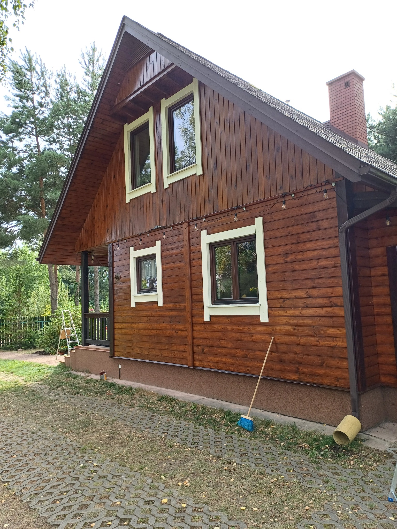 Drewniany dom jednorodzinny z brązową elewacją po renowacji, widoczne odświeżone ramy okienne i dekoracyjne lampki, na ziemi stoi miotła.