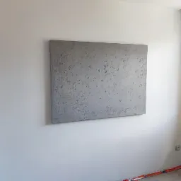 Szara ściana z efektem dekoracyjnym betonu architektonicznego, widoczna taśma malarska przy podłodze i gniazdko elektryczne na białej ścianie.