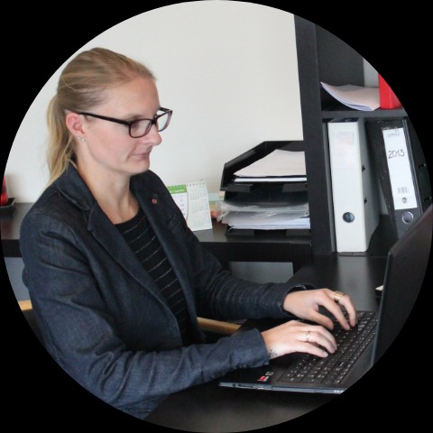 Kobieta w okularach i marynarce pracuje na laptopie przy biurku, widoczne segregatory i kalendarz.