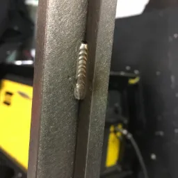 Spoina spawalnicza na metalowym profilu, widoczne ślady po spawaniu, w tle żółty sprzęt spawalniczy.
