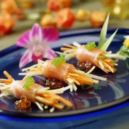 Elegancko podane roladki z łososia wypełnione julienne z warzyw, udekorowane kawiorem i kwiatami na niebieskim talerzu, w tle dekoracje kwiatowe.