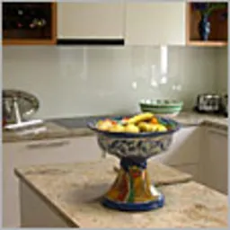 Szklana ściana między blatem a szafkami kuchennymi, odbijająca światło, z dekoracyjną misą na blacie. Widoczne fragmenty szafek w jasnym kolorze i inne elementy wyposażenia kuchni.