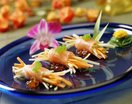 Elegancko podane roladki z łososia wypełnione julienne z warzyw, udekorowane kawiorem i kwiatami na niebieskim talerzu, w tle dekoracje kwiatowe.