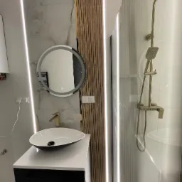 Nowoczesna łazienka z okrągłym lustrem z podświetleniem LED, złotą armaturą i kabiną prysznicową ze złotym zestawem natryskowym. Eleganckie wykończenie wnętrz.