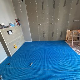 Apart Renovation Mateusz Dach - Niebieska mata pod ogrzewanie podłogowe w trakcie montażu, ściany wyłożone płytami Ultrament, drabina w tle. Etap prac przed wylaniem posadzki.