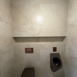 Apart Renovation Mateusz Dach - Nowoczesna łazienka z beżowymi płytkami imitującymi marmur, wiszącą toaletą i pisuaru w kolorze grafitowym. Minimalistyczny design z wnęką i ukrytymi przyciskami.