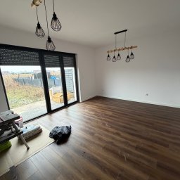 Apart Renovation Mateusz Dach - Jasne wnętrze z panelami podłogowymi i nowoczesnym oświetleniem. Widoczne okno z roletami zewnętrznymi i elementy przygotowania do prac wykończeniowych.