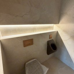 Apart Renovation Mateusz Dach - Nowoczesna toaleta w beżowych płytkach z podświetleniem LED, wiszącym bidetem i ukrytą spłuczką. Częściowo zakryta miska WC folią ochronną.