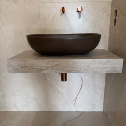 Apart Renovation Mateusz Dach - Beżowa łazienka z umywalką w kształcie misy na kamiennym blacie, miedziana armatura i płytki imitujące trawertyn. Minimalistyczny design z widocznymi przewodami.