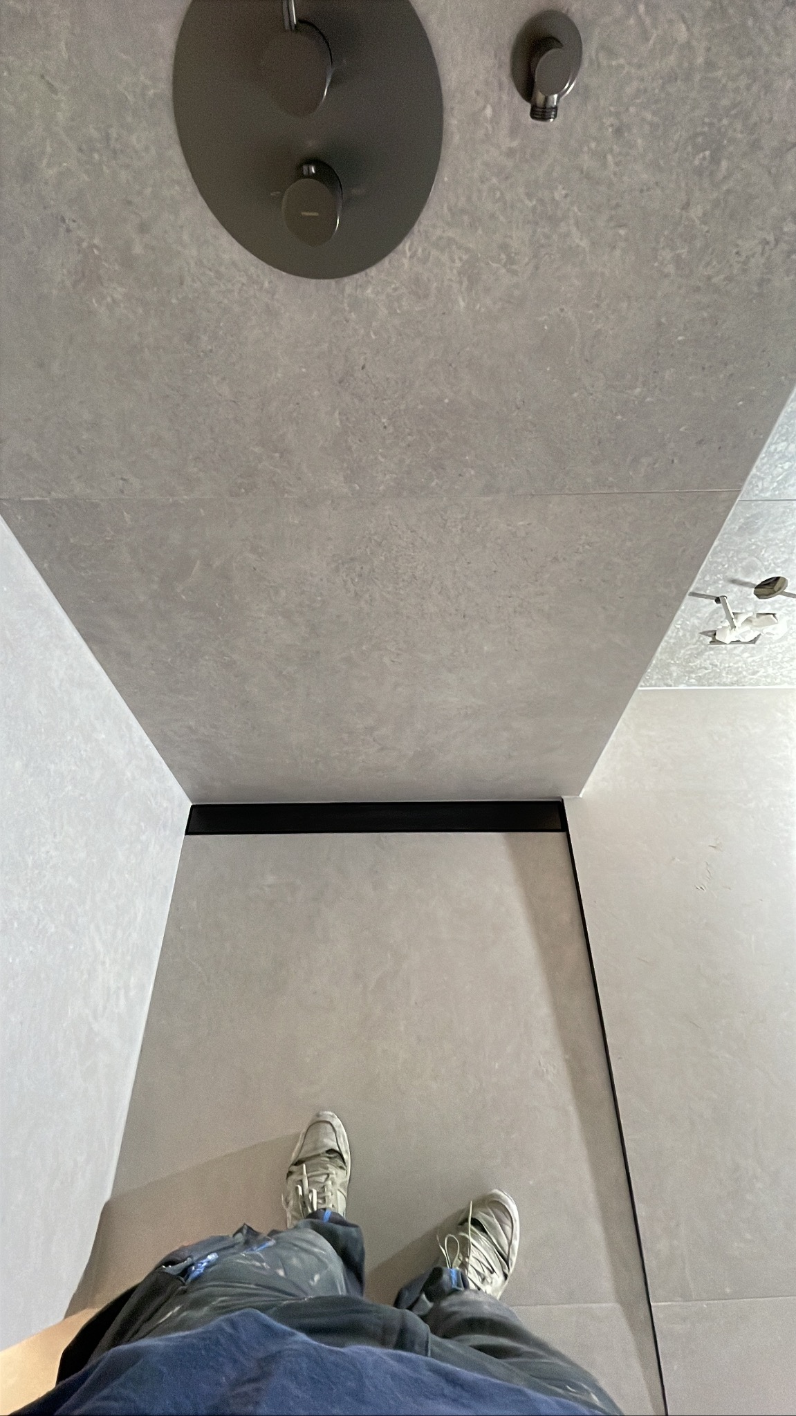 Nowoczesna kabina prysznicowa z szarymi płytkami, widok z góry na deszczownicę i odpływ liniowy. Drobne zabrudzenia na spodniach osoby robiącej zdjęcie.