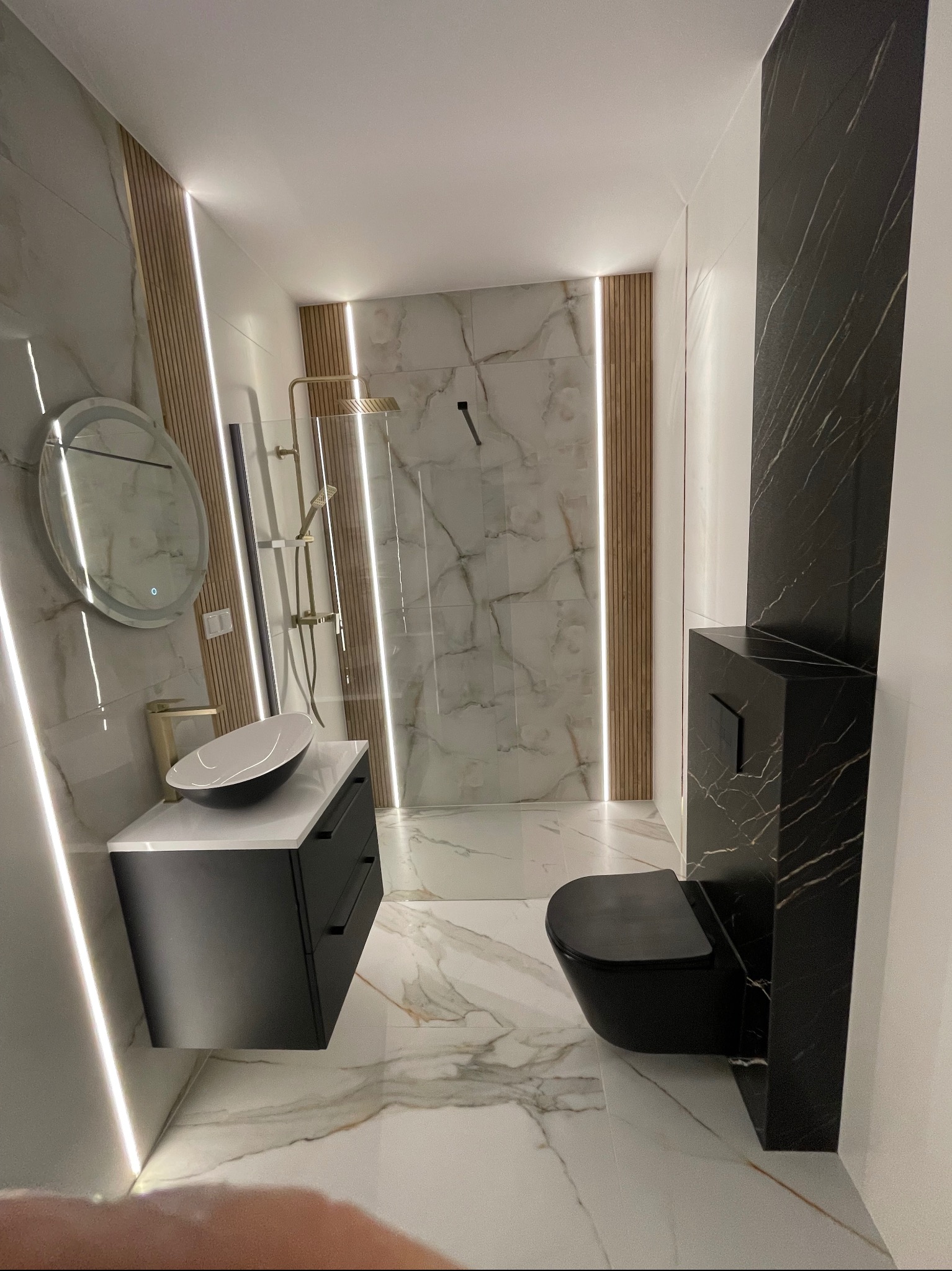 Nowoczesna łazienka z czarną armaturą i marmurowymi płytkami. Umywalka nablatowa, podświetlane listwy LED. Kabina prysznicowa z deszczownicą. Minimalistyczny design.