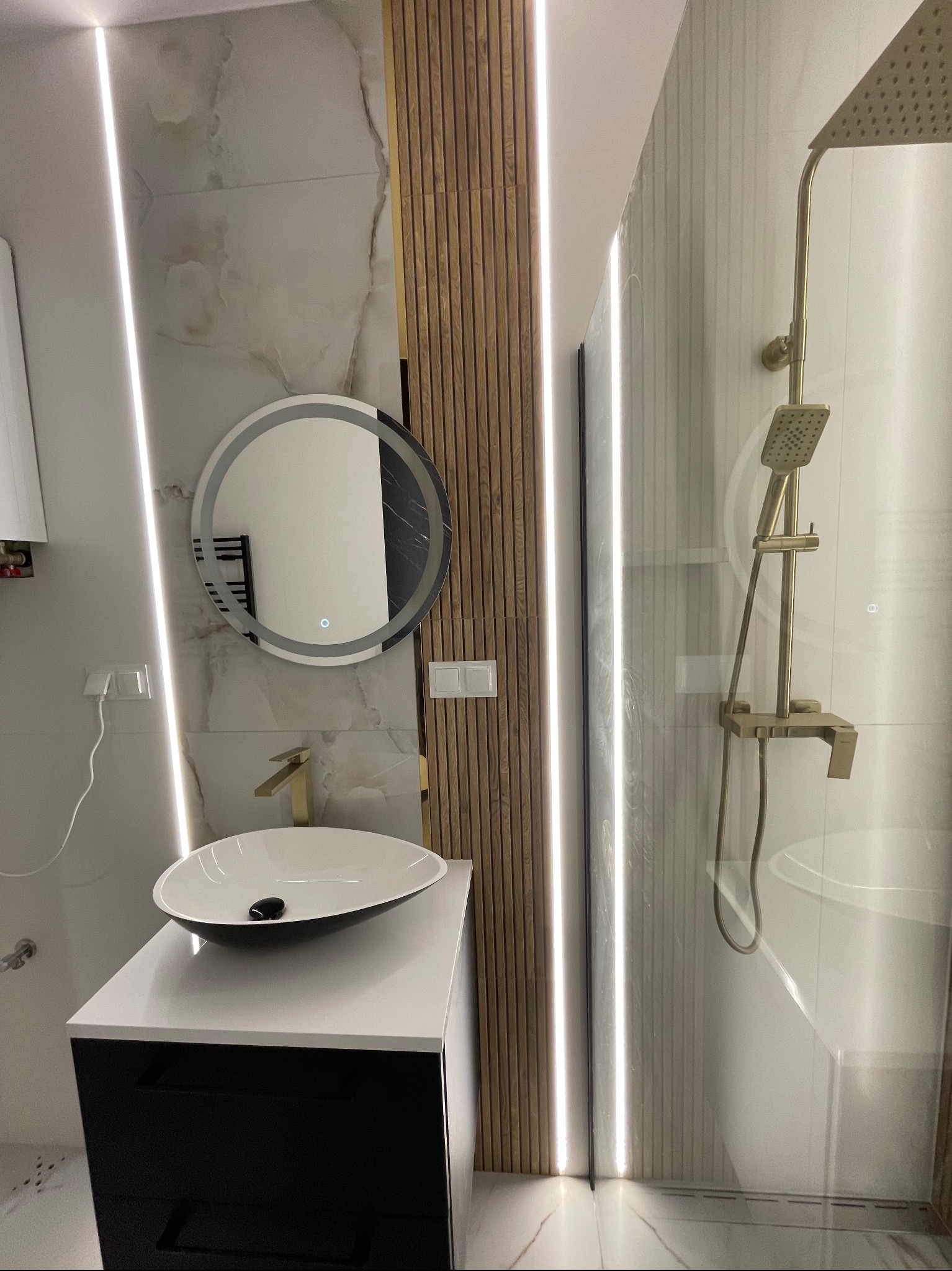 Nowoczesna łazienka z okrągłym lustrem z podświetleniem LED, złotą armaturą i kabiną prysznicową ze złotym zestawem natryskowym. Eleganckie wykończenie wnętrz.